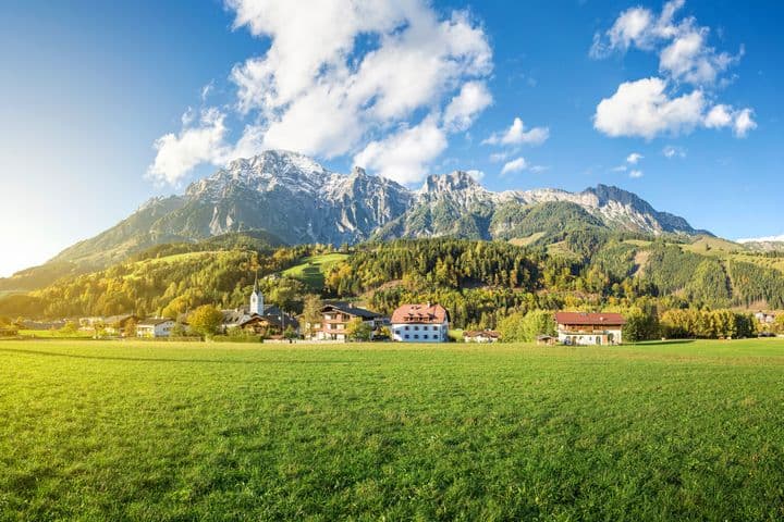 Landschaft in Leogang mit grünen Wiesen, einer Kirche, traditionellen Häusern und imposanten Bergen im Hintergrund bei klarem Himmel.