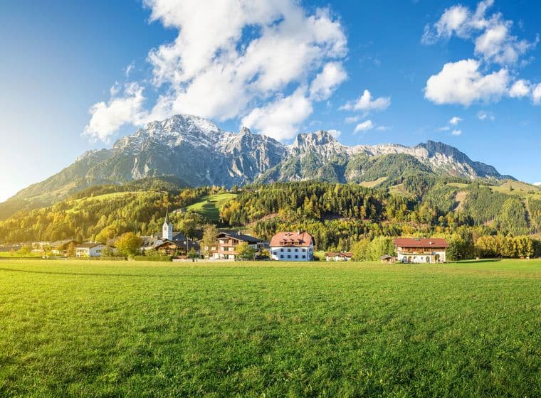Landschaft in Leogang mit grünen Wiesen, einer Kirche, traditionellen Häusern und imposanten Bergen im Hintergrund bei klarem Himmel.