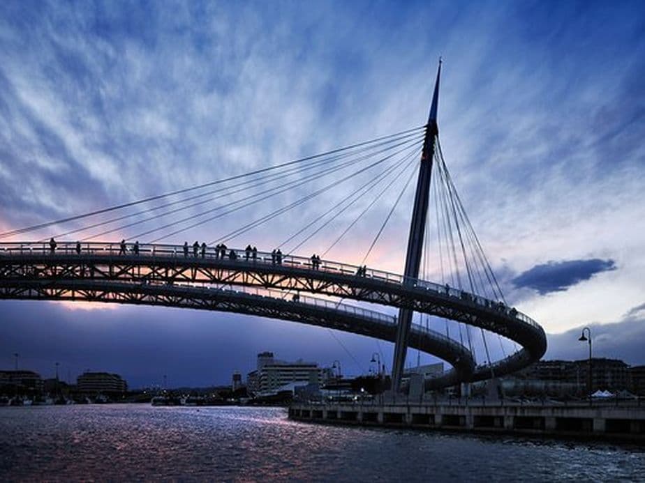 Silhouetted footbridge with cables arches over a river at dusk, with a dramatic cloudy sky and soft twilight hues in the background.