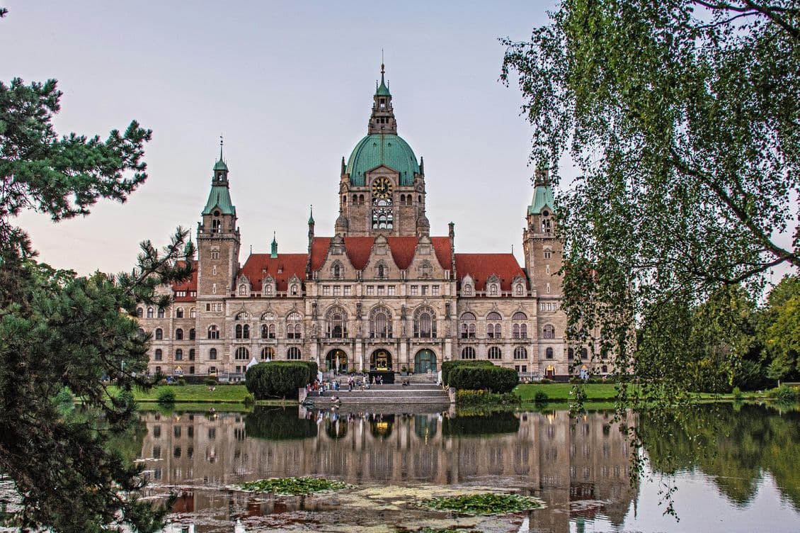 Historic building with a red roof and green domes, reflected in a tranquil pond, framed by trees on both sides under a clear sky.