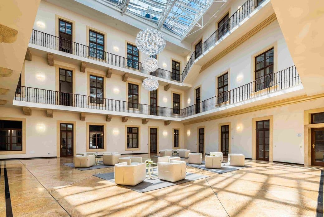 Interior of a bright atrium with a glass ceiling, designer chandeliers, and seating with white armchairs. Three floors of the building feature evenly spaced doors and balcony railings, creating a harmonious and airy space.