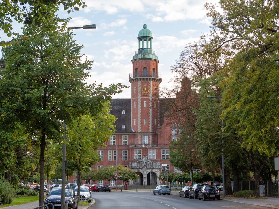Reinickendorf city hall, green-domed tower, surrounded by trees and a street with parked cars in the foreground.