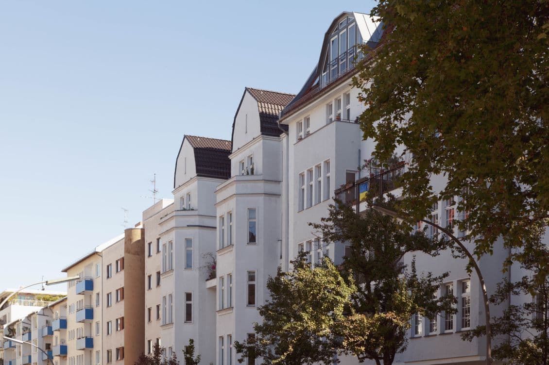 Row of elegant, multi-story residential buildings with white facades and gabled roofs, lined with trees under a clear blue sky.
