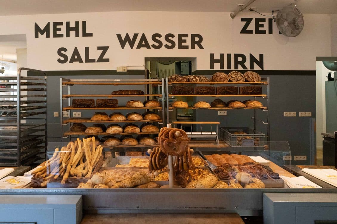A bakery display with various breads and pastries, including pretzels and loaves, under signs reading "Mehl," "Salz," "Wasser," and "Hirn."