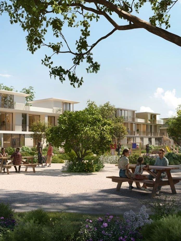 People sitting at picnic tables in a sunny park with modern apartment buildings and greenery in the background.