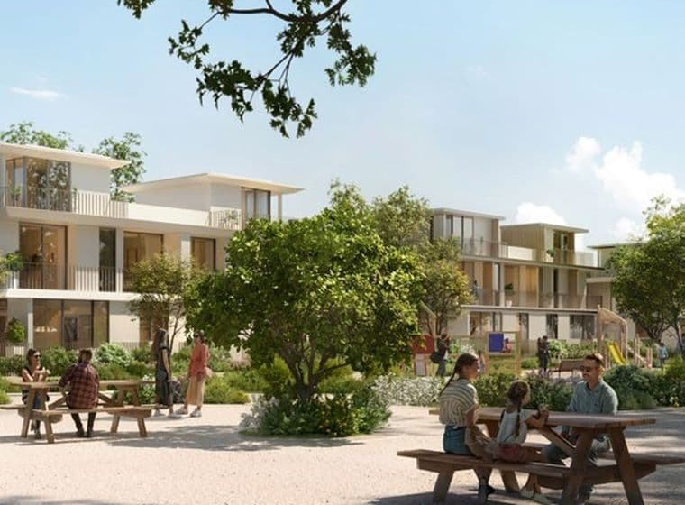 People sitting at picnic tables in a sunny park with modern apartment buildings and greenery in the background.