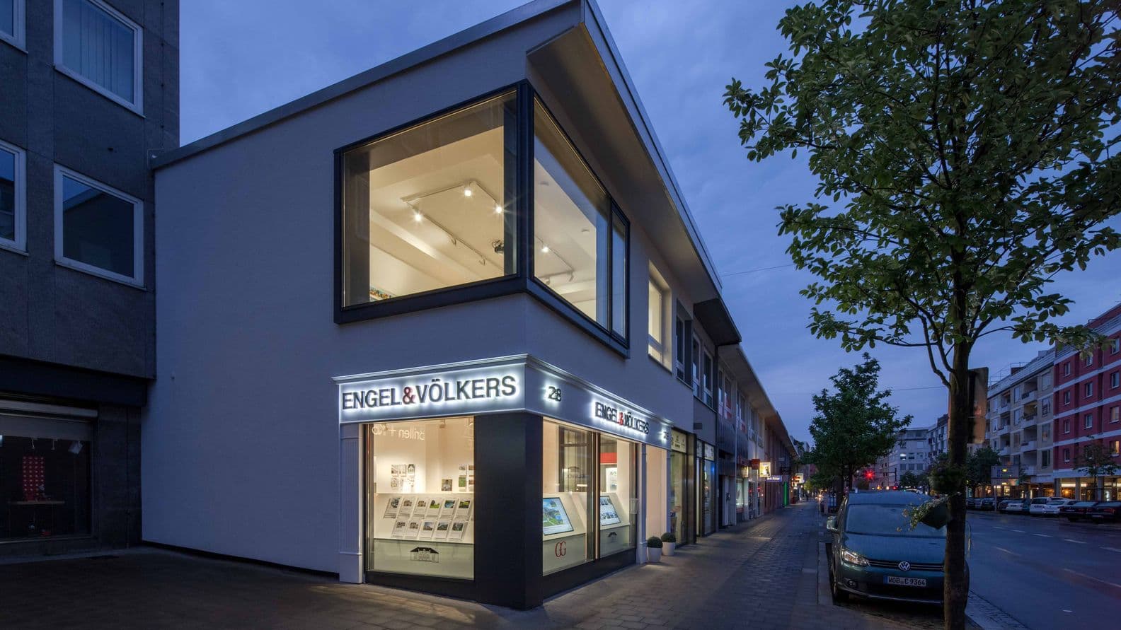 Modernes Gebäude mit großen Fenstern und „ENGEL & VÖLKERS“-Schriftzug, beleuchtet in der Abenddämmerung. Straßenansicht mit geparkten Autos und Bäumen.
