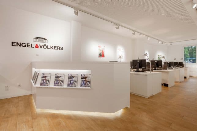 Modernes Immobilienbüro mit weißen Wänden, Holzfußboden, Schreibtischen mit Computern, ausliegenden Broschüren und großen Fenstern.