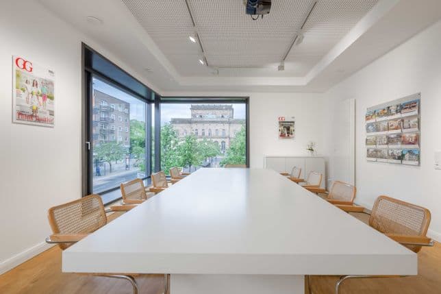 Moderner Konferenzraum mit einem langen weißen Tisch, Holzstühlen, großen Fenstern und Blick auf die Stadt. An den Wänden hängen bunte Kunstwerke und Zeitschriften.