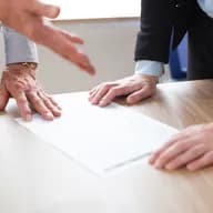 Two people leaning over a wooden table with a document: one has a hand resting on the paper, while the other reaches towards it.