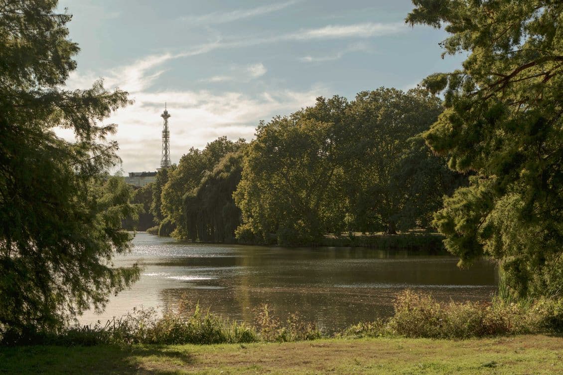 A serene lake surrounded by lush trees under a partly cloudy sky, with a distant tower visible in the background.