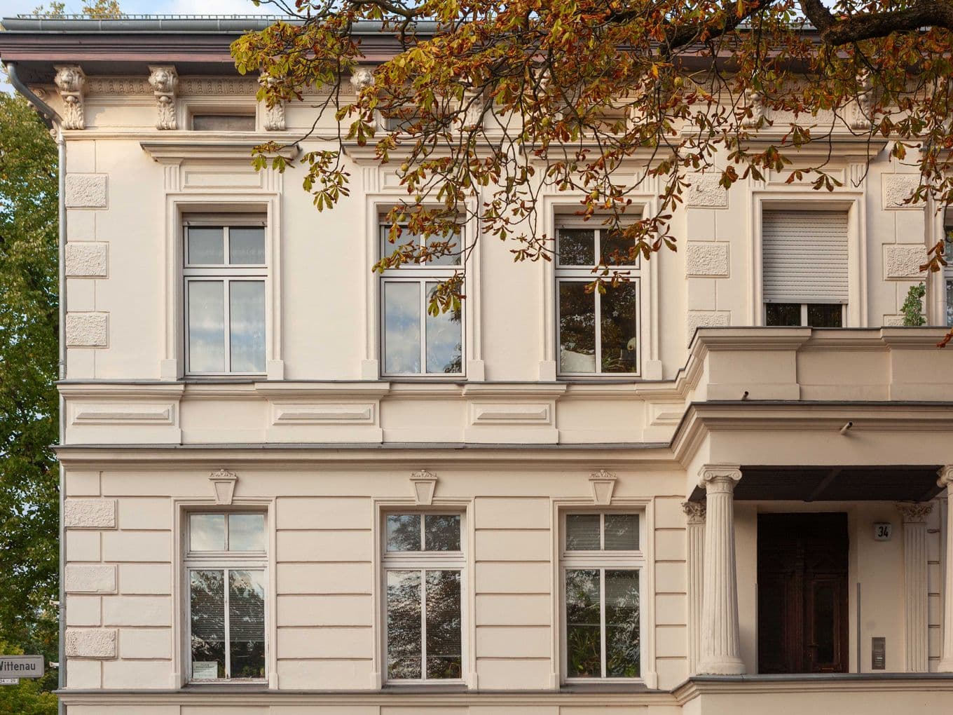 Elegant white building with large windows, ornate details, and a small balcony. Autumn leaves partially frame the scene.