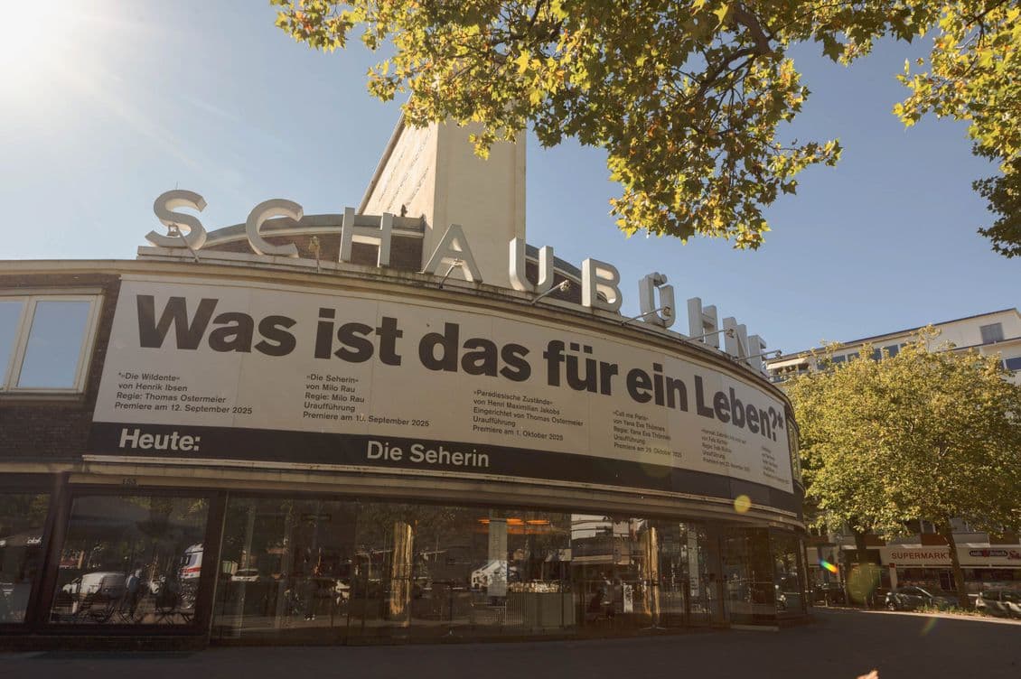 Theater with a large banner displaying "Was ist das für ein Leben?" under a clear sky and surrounded by trees.