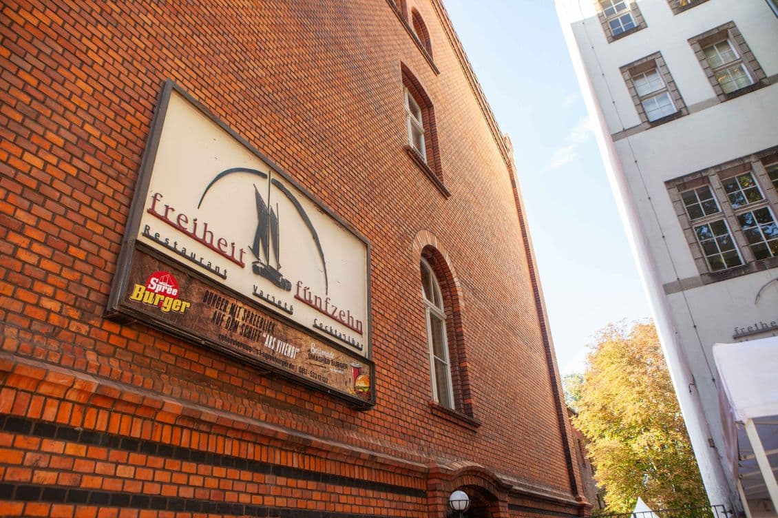 Brick building with a large sign reading "Freiheit Fünfzehn" and a smaller "Spree Burger" logo. Trees and another building in the background.