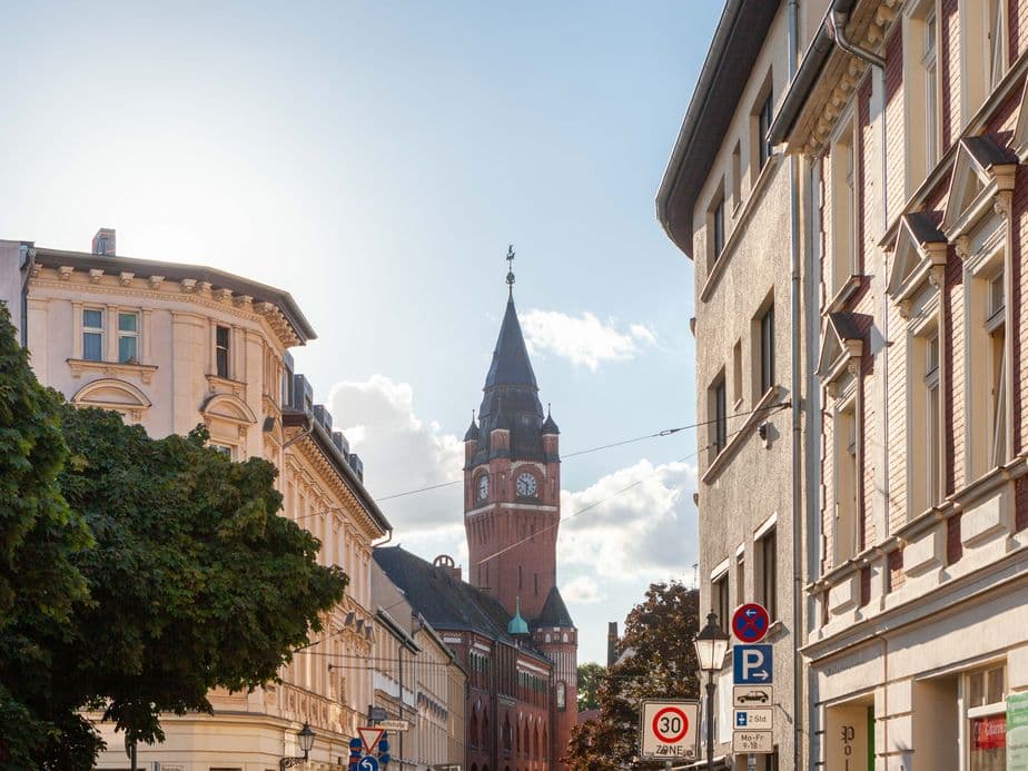Street view with historic buildings and a tall clock tower under a clear sky, trees lining the street, and parked cars visible.