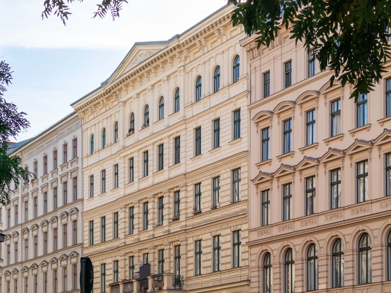 Historic European-style buildings with ornate facades and arched windows under a clear sky, framed by tree branches.