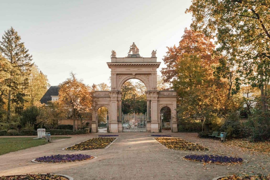 Ornate stone archway in a park during autumn, surrounded by colorful trees and flowerbeds under a clear sky.