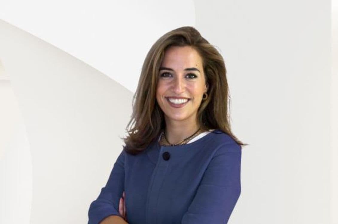 Eine lächelnde Frau mit braunem Haar, die eine blaue Jacke trägt, steht vor einem weißen Hintergrund.