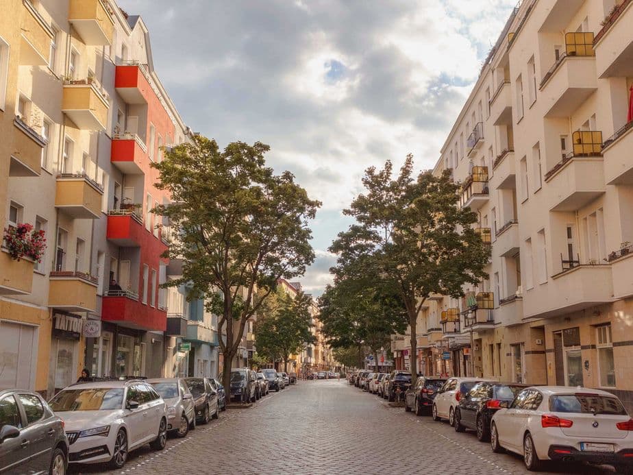 A cobblestone street lined with parked cars and trees, flanked by colorful apartment buildings under a partly cloudy sky.