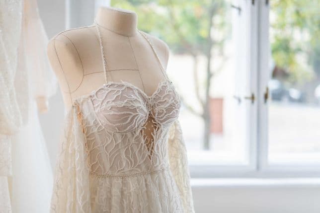 Close-up of a lace wedding dress on a mannequin by a window. Blurred background shows a tree and a street outside.