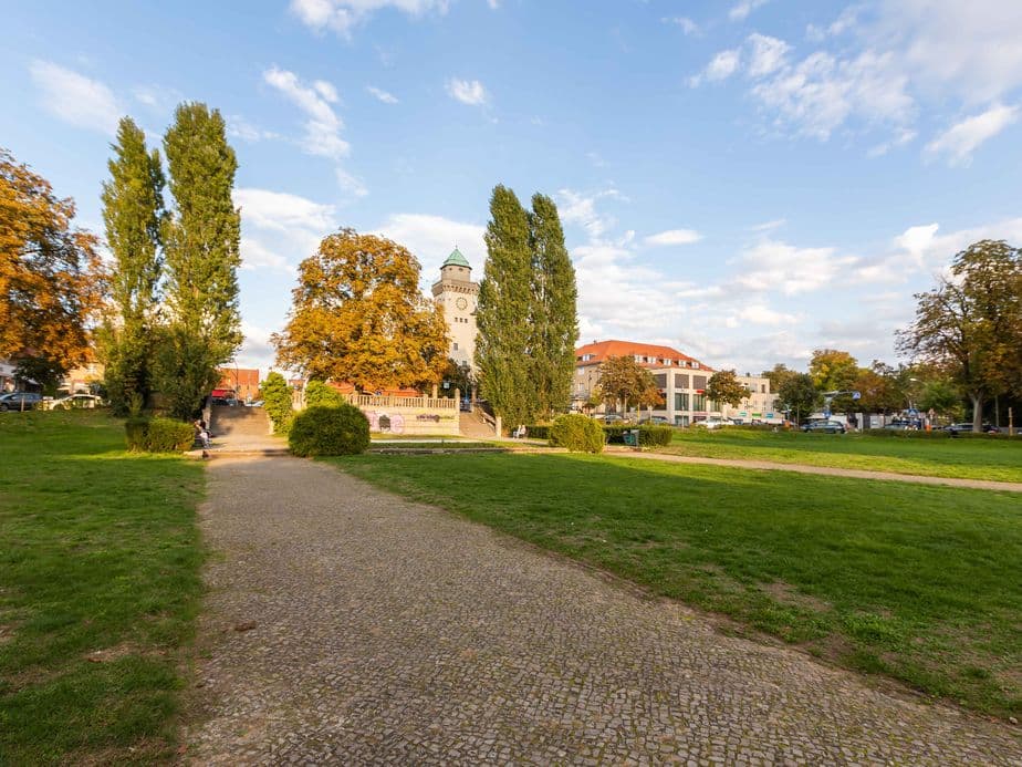 A park with a cobblestone path, tall trees, and a clock tower in the background under a partly cloudy sky.