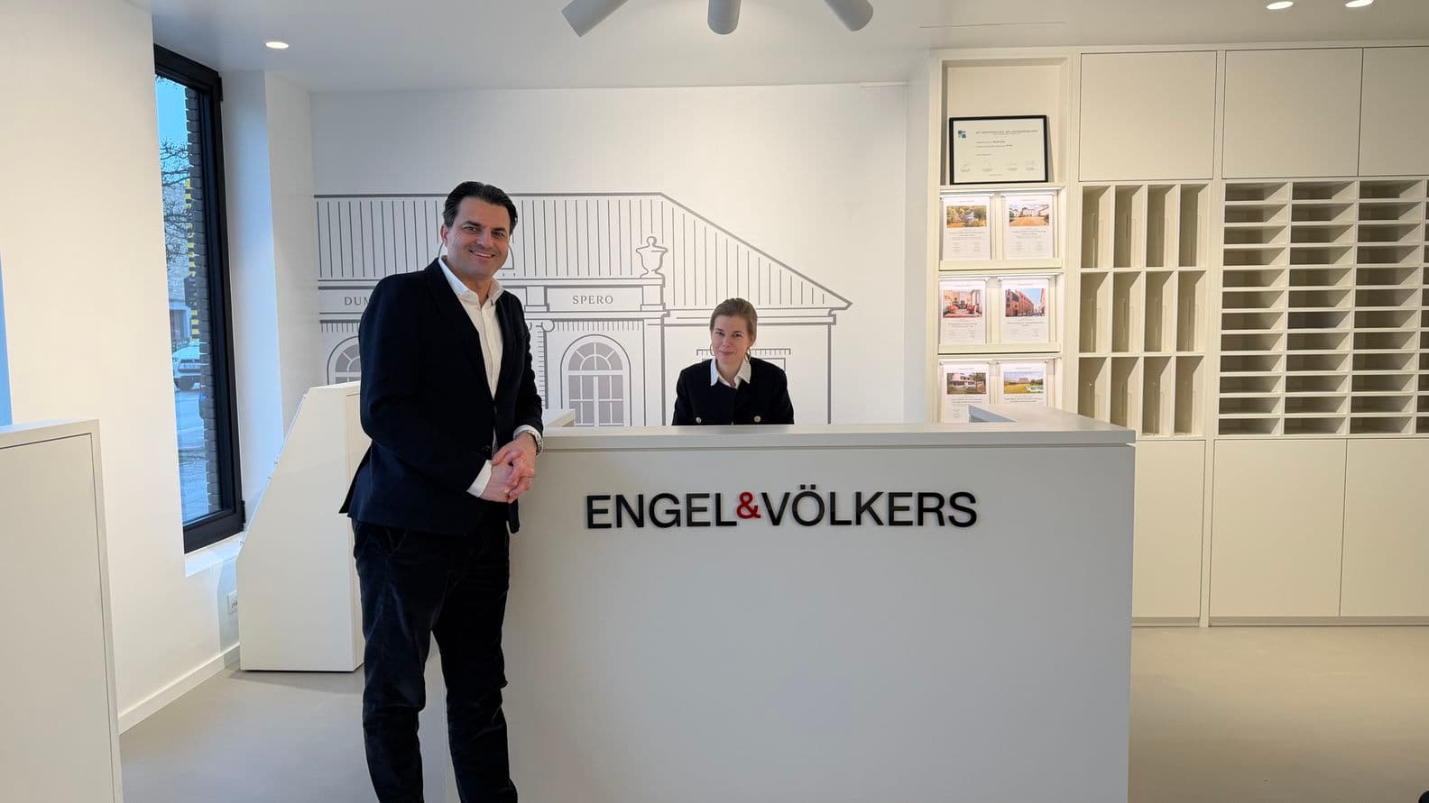 A man stands at a white reception desk labeled "Engel & Völkers," speaking with a receptionist in a modern, well-lit office.