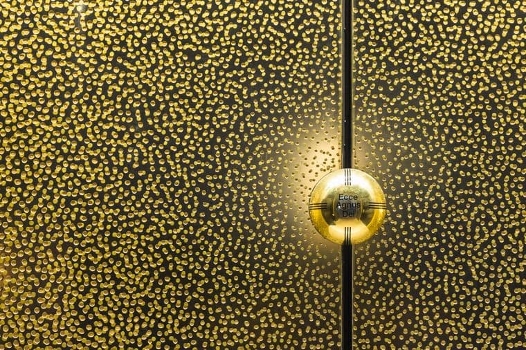 Close-up of a gold door with small circular cutouts and a round door handle with the inscription "Ecce Agnus Dei".