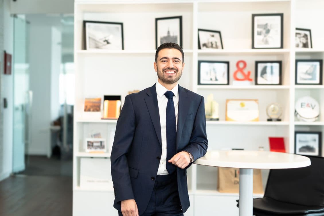 Smiling man in navy suit and tie leaning on a round table in a bright office with shelves of framed photos and awards.