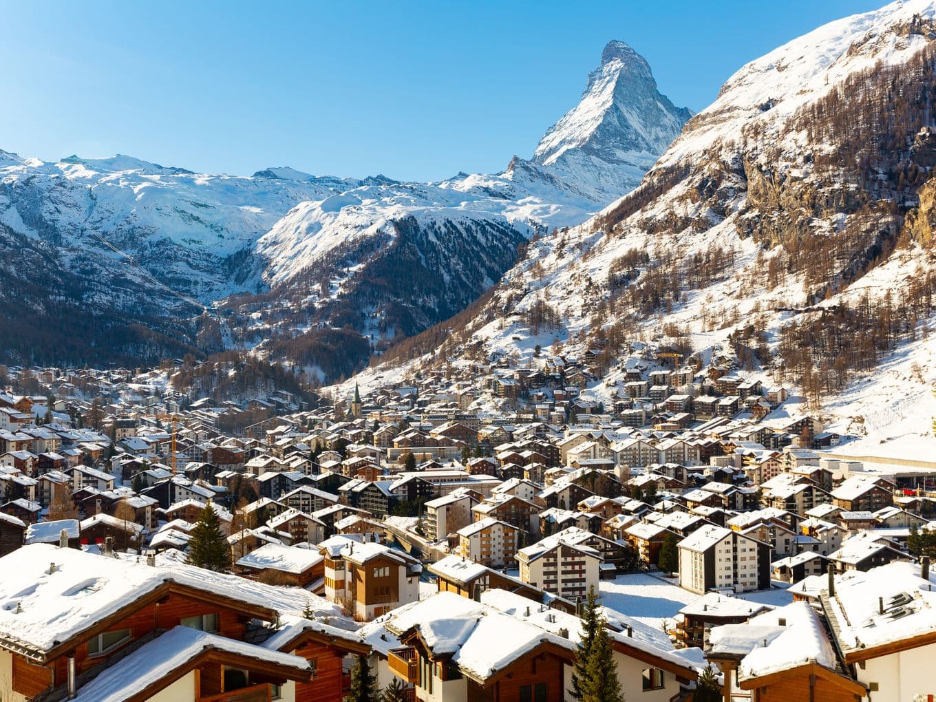 Winterliche Panoramaaufnahme des Schweizer Bergdorfs Zermatt mit schneebedeckten Chalets im Vordergrund und dem markanten Matterhorn im Hintergrund unter blauem Himmel.