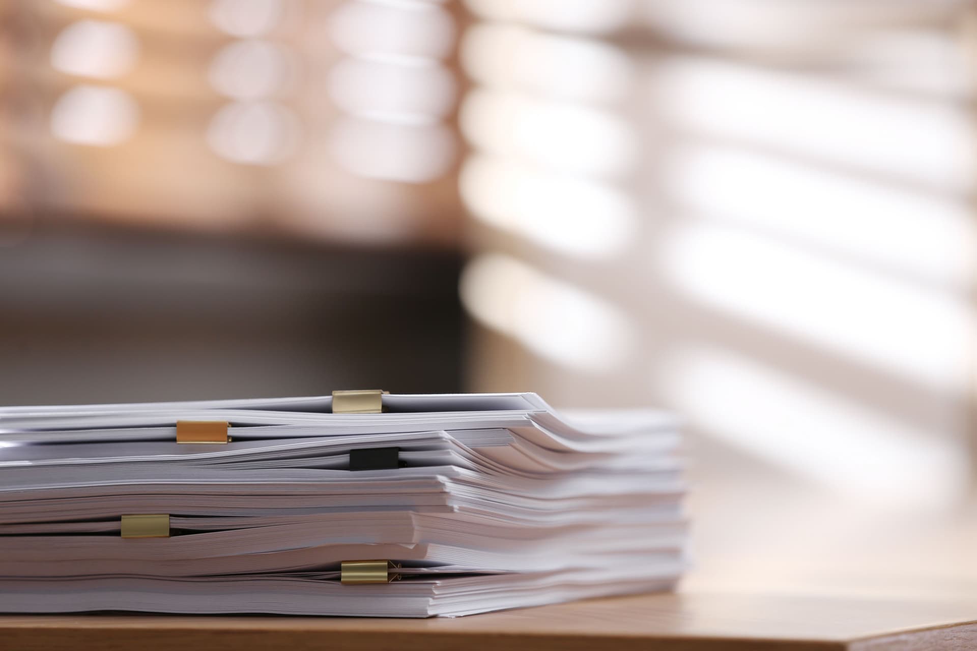 A stack of documents with paper clips on a wooden desk, illuminated by soft, diffused sunlight through blinds in the background.