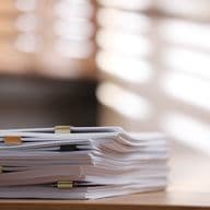 A stack of documents with paper clips on a wooden desk, illuminated by soft, diffused sunlight through blinds in the background.