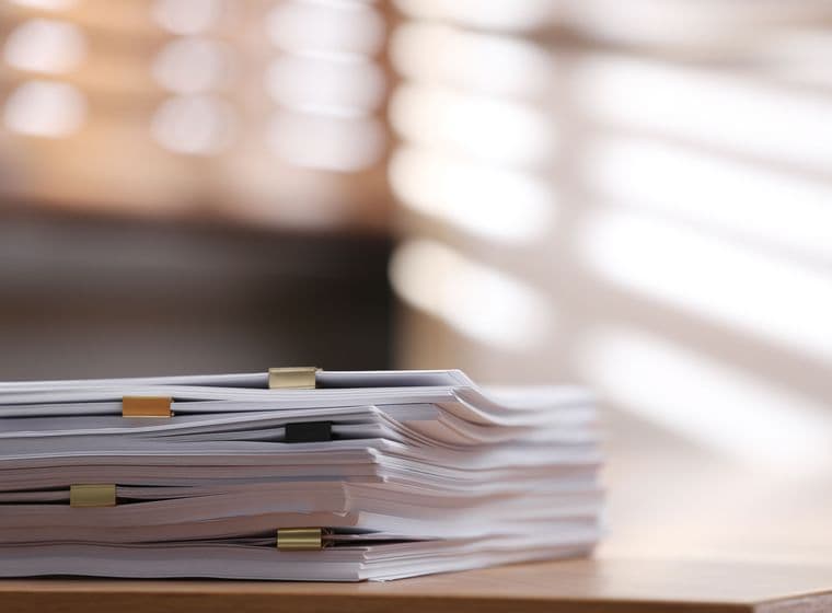 A stack of documents with paper clips on a wooden desk, illuminated by soft, diffused sunlight through blinds in the background.