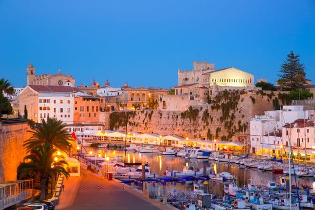 Vista iluminada del pintoresco puerto de Ciutadella, Menorca, de noche: reflejos que bailan sobre el agua y edificios históricos brillando en la atmósfera vespertina.