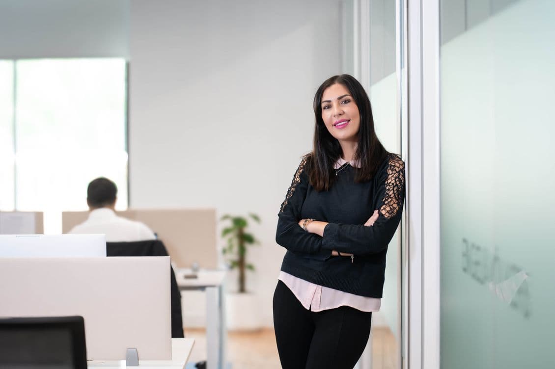 Woman standing with arms crossed, smiling in a modern office near a glass partition. Blurred coworkers are visible in the background.