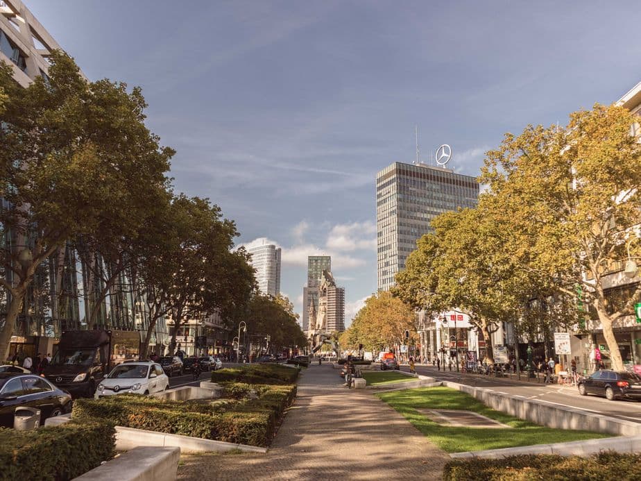 Urban street scene with tall buildings, trees lining the road, parked cars, and people walking on a path under a clear blue sky.
