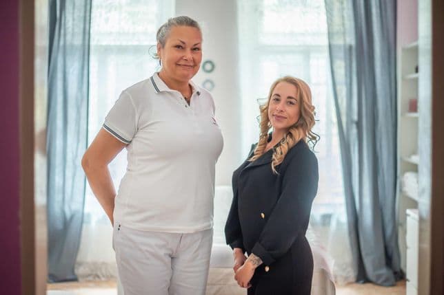 Two women standing side by side in the studio interior. Šárka Eggerová on the left in a white polo shirt, Eliška Tichá on the right in a black coat. Soft light filters through curtains and windows in the background.