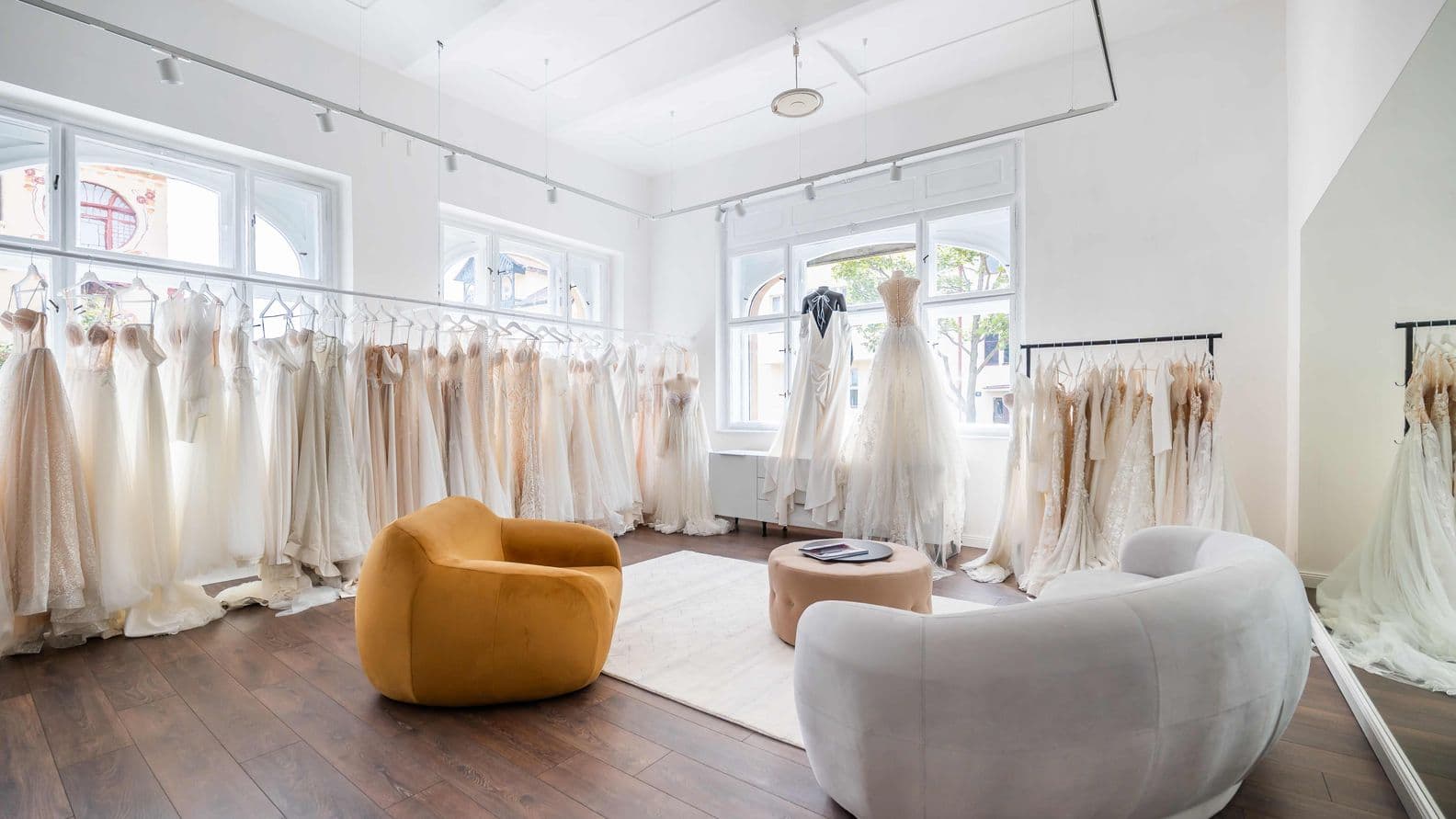 Interior of a bridal salon with rows of wedding dresses on hangers, large white windows, and modern seating. A mustard armchair on the left, white armchair on the right, round coffee table in the center on wooden flooring.