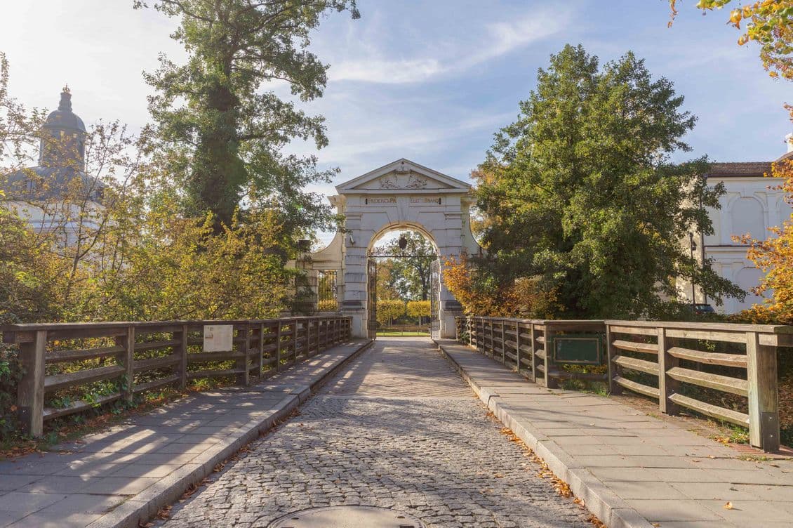 Stone archway on a cobblestone path flanked by trees and railings, under a clear sky.