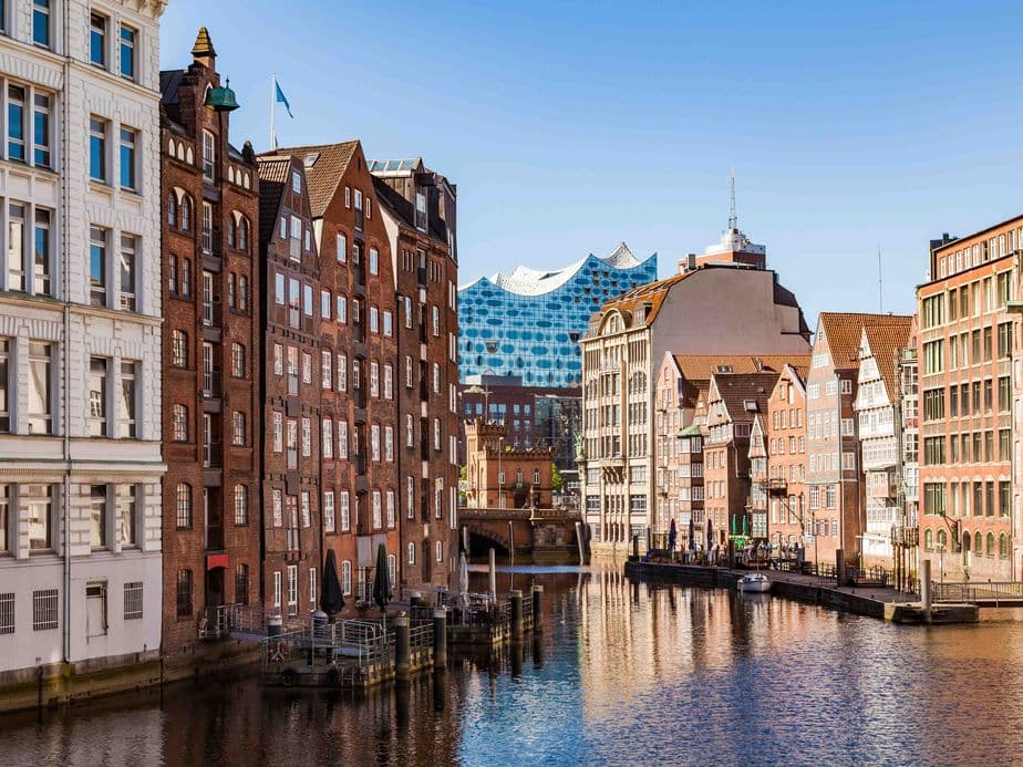 A scenic canal in Hamburg, lined with historic brick buildings, under a clear blue sky, with the Elbphilharmonie in the background.