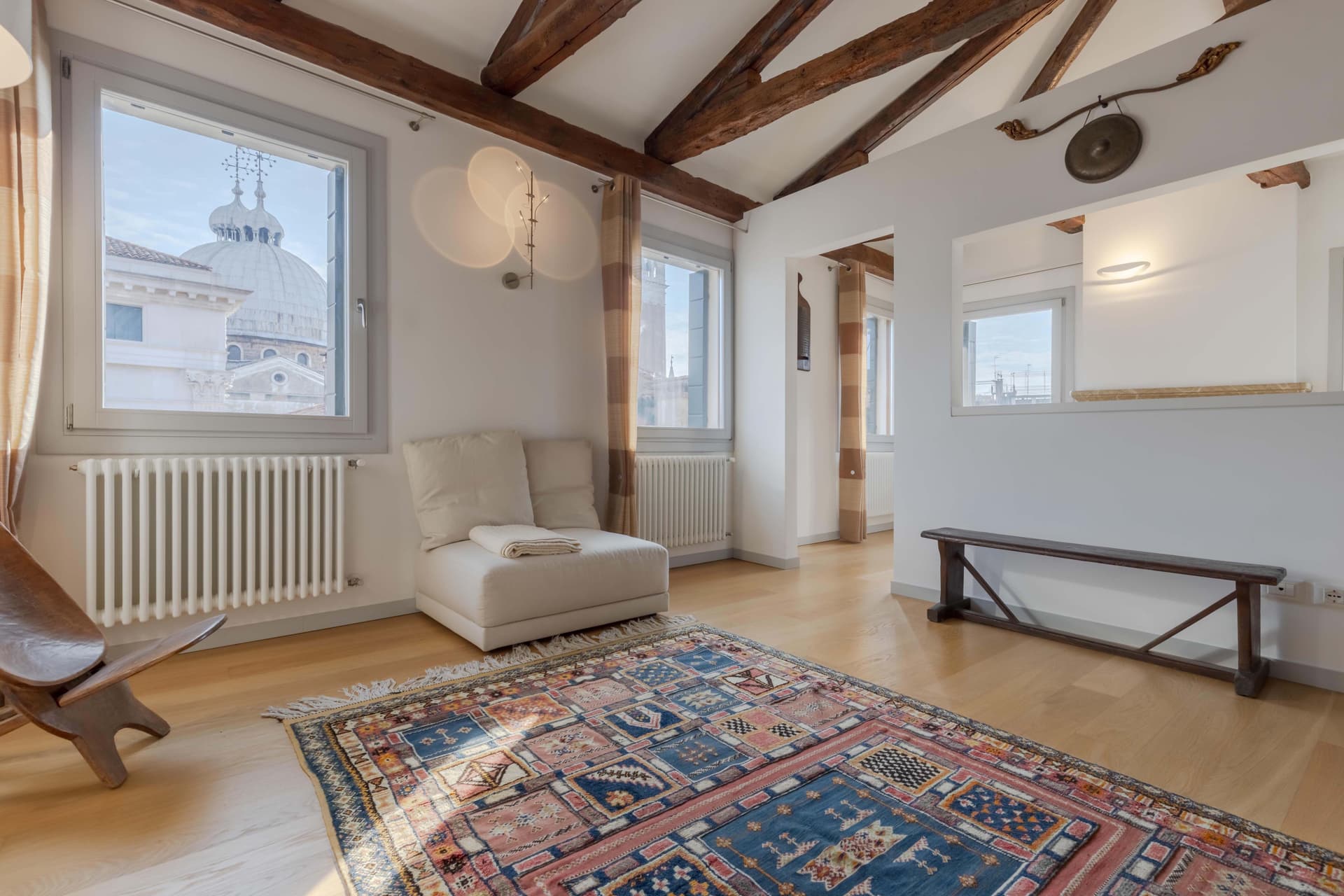 The interior of an elegant Venetian apartment with exposed wooden beams on the white ceiling. The bright room features white walls, light wood flooring, and a window offering a stunning view of a Venetian church dome. The furnishings include a white armchair, an antique Persian rug in shades of blue and pink, a radiator beneath the window, and minimalist décor elements. A small decorative gong is also hung on the wall. The space blends traditional Venetian elements with contemporary style.