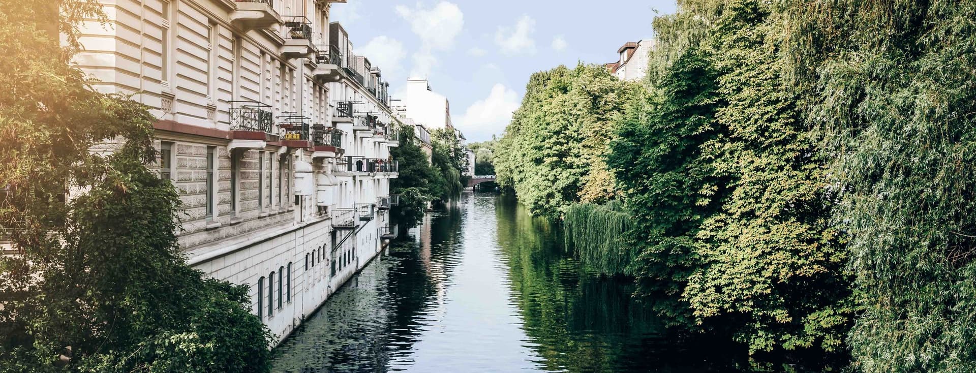 A tranquil canal, lined with lush green trees in Eppendorf and classic European buildings under a clear blue sky.