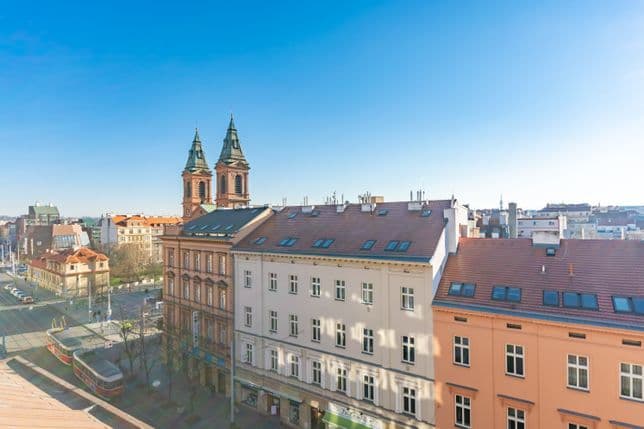 Pohled z výšky na pražské ulice s historickými domy, dvěma věžemi kostela, projíždějící tramvají a modrou oblohou.
