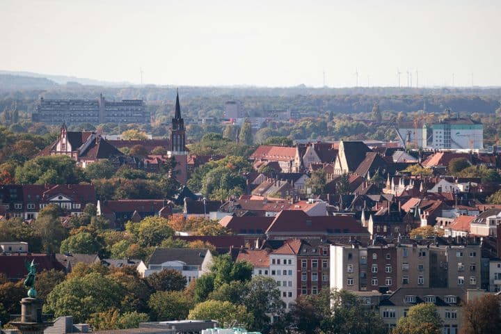 Aerial view of a Hannover with red-roofed buildings, a prominent church tower, and surrounding trees under a clear sky.