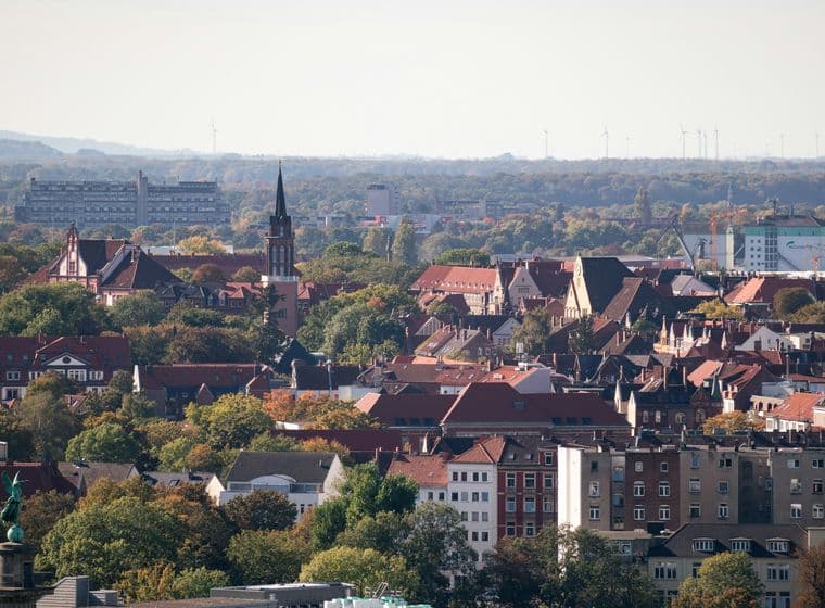 Aerial view of a Hannover with red-roofed buildings, a prominent church tower, and surrounding trees under a clear sky.