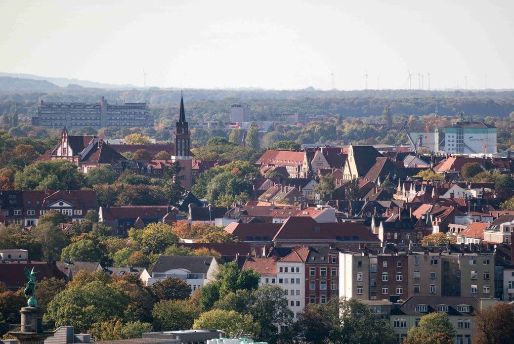 Aerial view of a cityscape with red-roofed buildings, a church steeple, and distant forested area under a hazy sky.