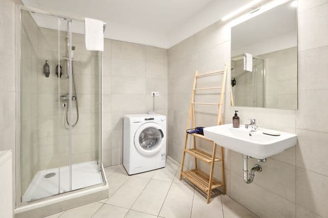 Bright bathroom finished with beige tiles. On the left there is a glass shower enclosure with a shower head and wall-mounted toiletry dispensers. Next to it stands a white washing machine. On the right side there is a wall-mounted sink with a tap and a large rectangular mirror with overhead lighting. Soap and a small dish are placed by the sink, and a wooden ladder-style shelf stands beside it. The floor is covered with light-coloured tiles.