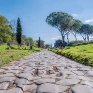 Cobblestone road lined with green trees under a blue sky. A cyclist rides in the distance.