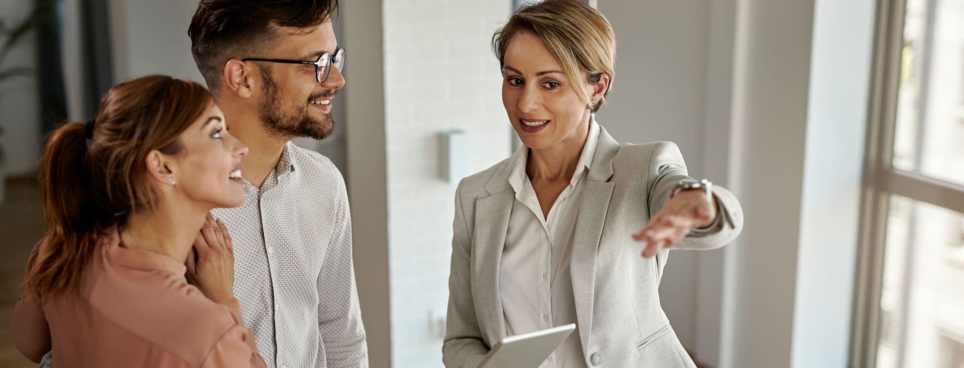 A real estate agent in a gray suit shows a young couple around a modern, bright apartment. She holds a tablet and gestures with her hand.