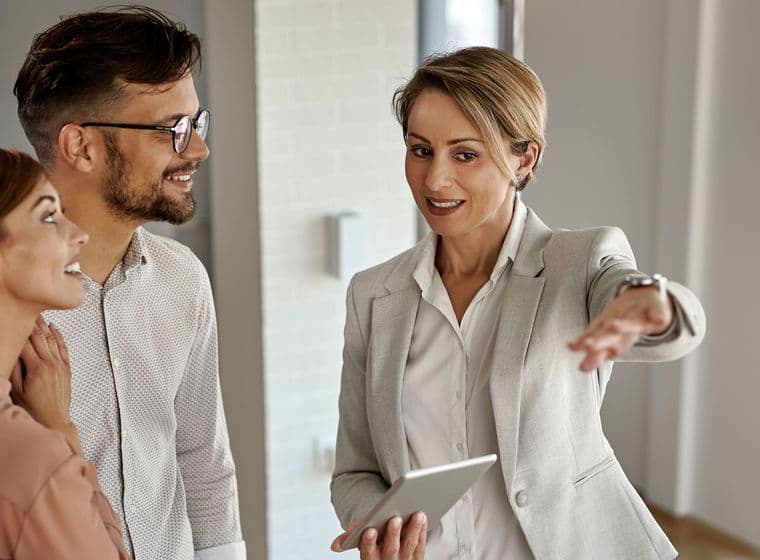 A real estate agent in a gray suit shows a young couple around a modern, bright apartment. She holds a tablet and gestures with her hand.