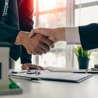 Two people in suits shake hands over a white desk with a house model, keys, and paperwork.